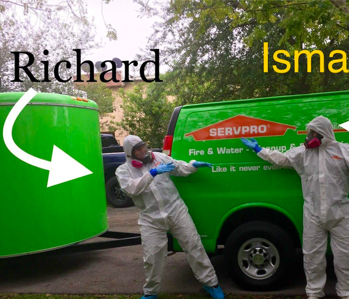 Employees in white protective suits pointing at each other in front of two green SERVPRO vans. 